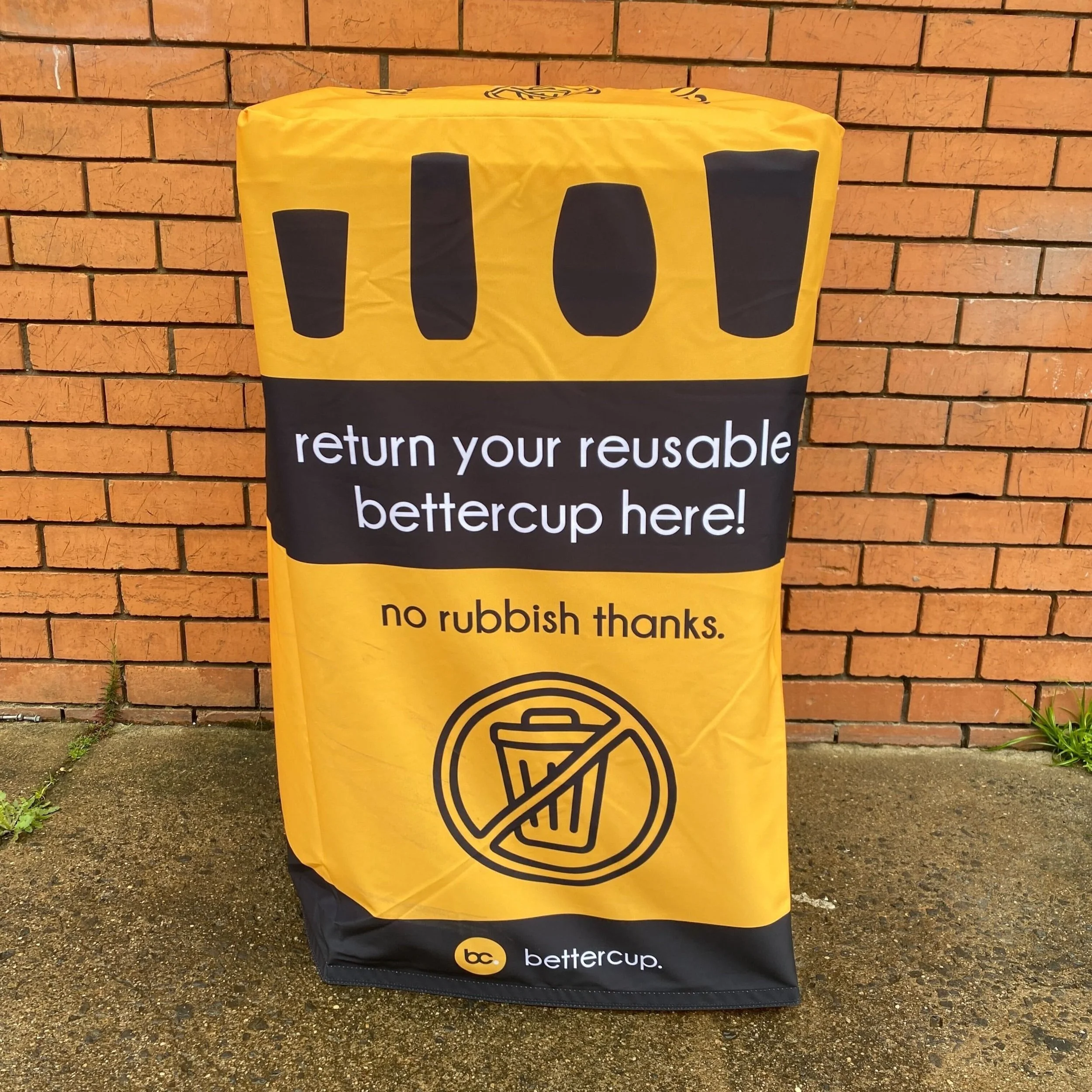 Orange and black printed fabric bin cover over a 240L bin with text that says "return your reusable bettercup here" and with a hole in the top for patrons to return their reusable cup.