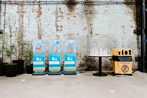 Three reusable cup dispensing stations and a bettercup bin set up against a brick wall, with a table of stacked bettercup tumblers nearby.
