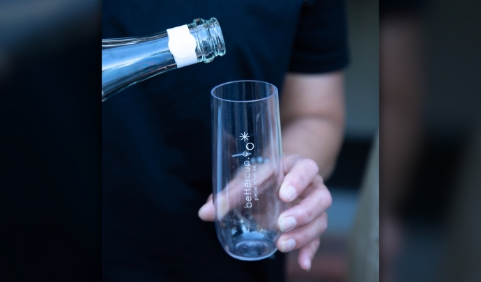 A person pouring a drink from a bottle into a bettercup champagne flute, held with a 'please return me' label visible.